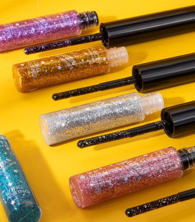 Glitter mascara tubes in purple, gold, silver, pink, and blue with black wands, arranged on a yellow background.