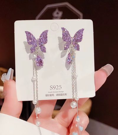 A pair of luxurious purple butterfly earrings with pearl accents, displayed on a white card and held by a hand with long nails.