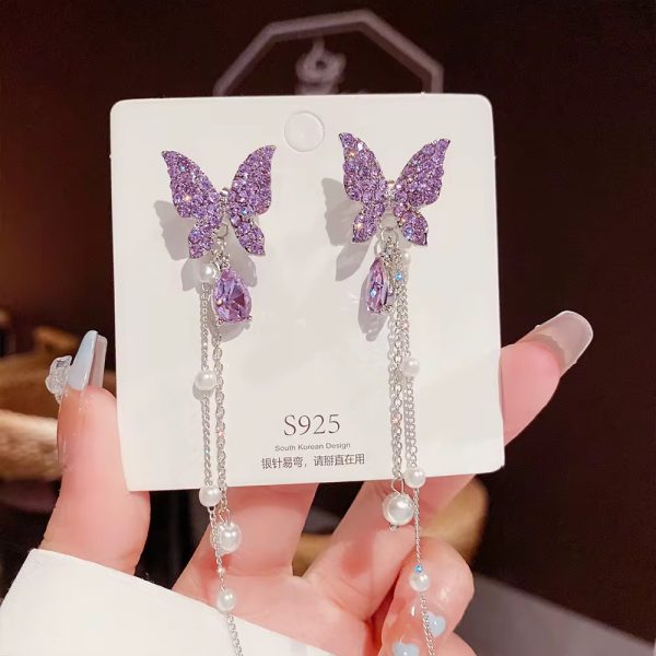 A pair of luxurious purple butterfly earrings with pearl accents, displayed on a white card and held by a hand with long nails.