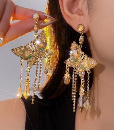 A close-up of a woman's ear wearing luxurious gold butterfly earrings with crystal dangling chains, showcasing the intricate details and sparkling crystals.