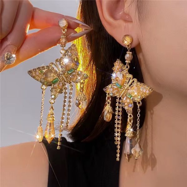 A close-up of a woman's ear wearing luxurious gold butterfly earrings with crystal dangling chains, showcasing the intricate details and sparkling crystals.