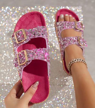A pair of pink rhinestone sandals with gold buckles on a sparkling background, held by a hand with glittery nails.