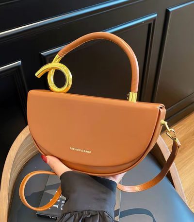 A luxurious brown half moon shoulder bag with gold accents, held by a woman's hand, showcasing its sleek design and sophisticated style.