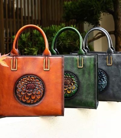 Three luxurious leather handbags in brown, green, and black, adorned with intricate floral embellishments, placed on a white surface amidst lush greenery.
