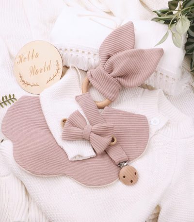 A beautifully arranged flat lay of a soft pink baby bib and pacifier clip set on a white knitted background, surrounded by lush greenery and a 'Hello World' wooden sign.