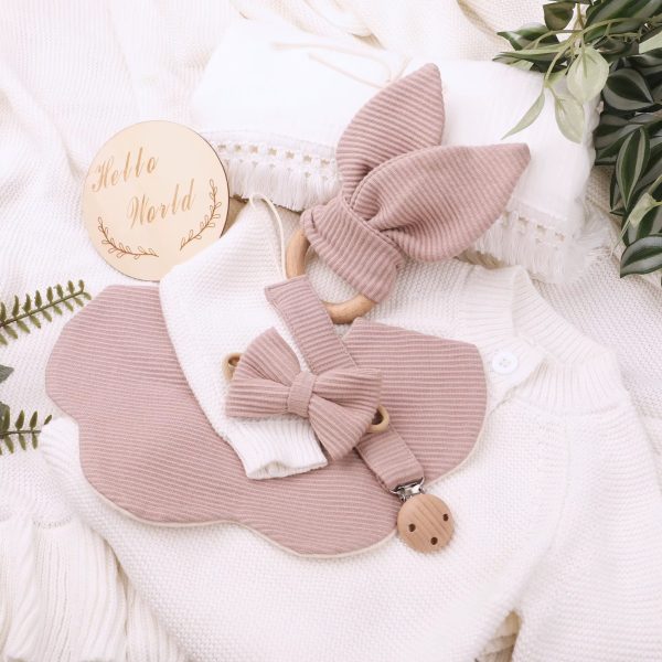 A beautifully arranged flat lay of a soft pink baby bib and pacifier clip set on a white knitted background, surrounded by lush greenery and a 'Hello World' wooden sign.