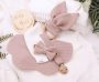 A beautifully arranged flat lay of a soft pink baby bib and pacifier clip set on a white knitted background, surrounded by lush greenery and a 'Hello World' wooden sign.